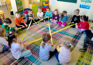 Dzieci siedząc na dywanie, tworzą ze wstążki "sieć internetową".
