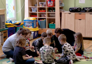 Dzieci siedzą z p.Olą i p. Martą na dywanie, chwytają klamerkami do bielizny , pomponiki z obręczy.