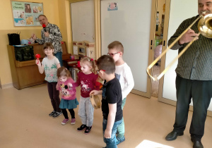 Dzieci utworzyły mała orkiestrę i grają na różnych instrumentach, w towarzystwie muzyka grającego na puzonie.