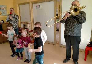 Dzieci utworzyły mała orkiestrę i grają na różnych instrumentach, w towarzystwie muzyka grającego na puzonie.
