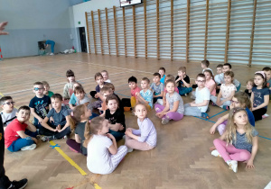 Dzieci siedzą w rozsypce w sali gimnastycznej.