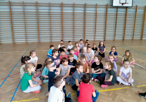 Dzieci siedzą w rozsypce w sali gimnastycznej.