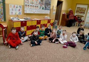 Dzieci siedzą na dywanie maja na twarzach karnawałowe maski.