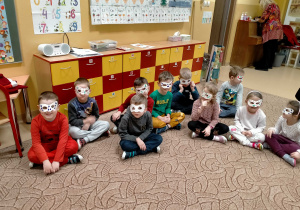 Dzieci siedzą na dywanie maja na twarzach karnawałowe maski.